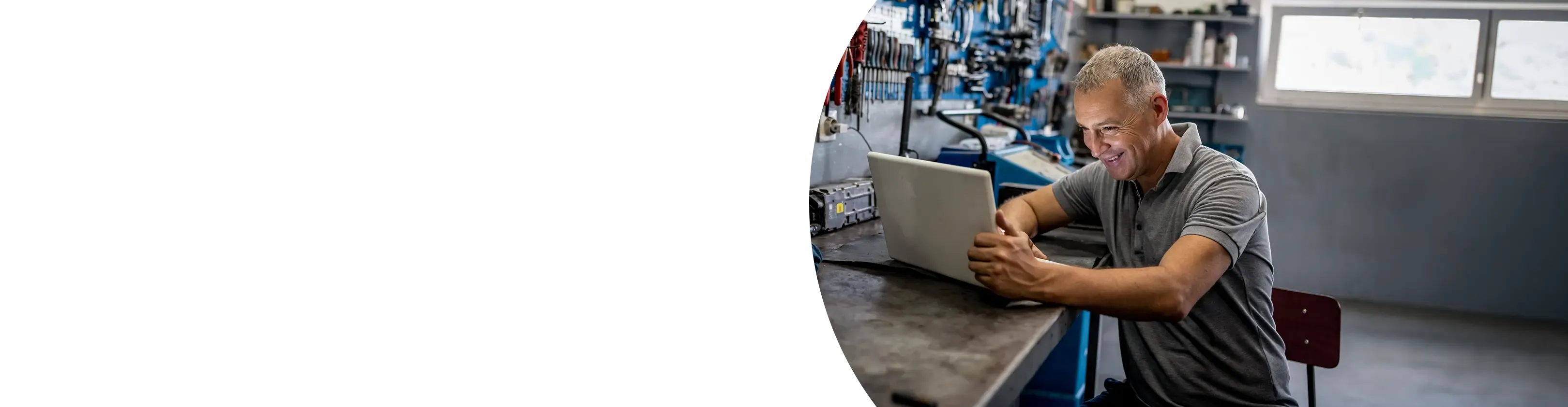 A smiling mechanic uses a laptop at his workbench in front of a pegboard of tools.